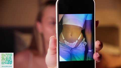 Video of a man holding a smartphone, displaying a close-up of a woman's midriff in a white crop top and blue jeans. The background is blurred, featuring a wooden table.