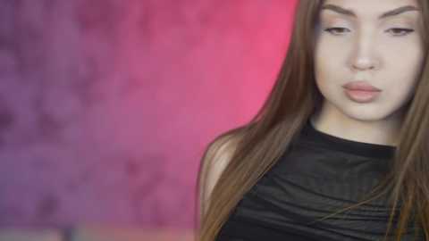 Video of a young East Asian woman with long, straight brown hair, wearing a black sleeveless top, standing against a blurred purple and pink background.
