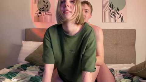 Video of a young woman with a short blonde bob wearing a loose green shirt, sitting on a man's lap in a modern bedroom with abstract wall art.