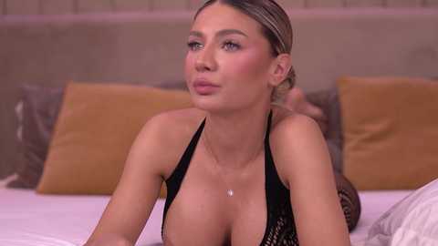 Video of a light-skinned woman with blonde hair in a ponytail, wearing a black lace halter top, lying on a bed with beige headboard and pillows.