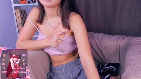 A video of an East Asian woman with straight, shoulder-length black hair, wearing a light pink bralette and gray sweatpants, sitting on a beige couch, pulling her bra strap.