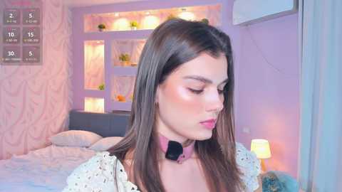 A video of a young woman with long brown hair, wearing a pink choker and white lace top, standing in a pastel-colored bedroom with a bed, shelves, and a lamp.