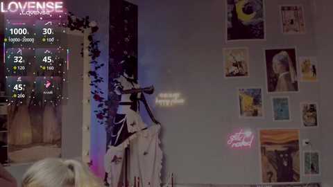 Video of a young woman with short, blonde hair, standing in a dimly lit room with a purple wall. She holds a guitar, surrounded by posters and a music chart displayed on a screen.