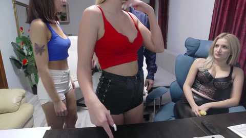 Media: Video of three women in a living room: a blonde in a red crop top and high-waisted shorts, a brunette in a blue top and white shorts, and a blonde in a black lace bra, all seated in blue armchairs.