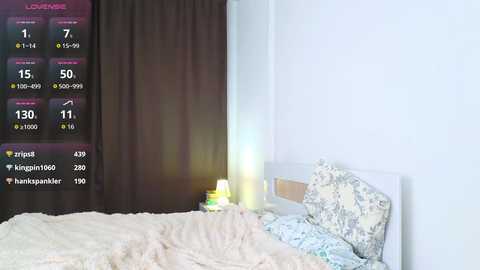 Video of a minimalist bedroom with a white bed, floral-patterned pillows, beige blanket, and dark curtains. Overlay displays health metrics: \"Loudness\" at 7, \"Sip\" at 1, \"Wrist\" at 7, \"Heart\" at 7, \"Breathing\" at 7, \"Temperature\" at 7.