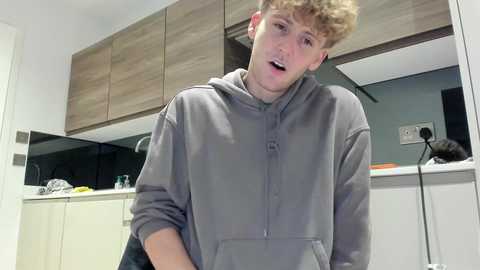 Video of a young, fair-skinned, blonde-haired man wearing a gray hoodie, standing in a modern kitchen with light wood cabinets and a white countertop. He appears surprised or shocked, mouth open.