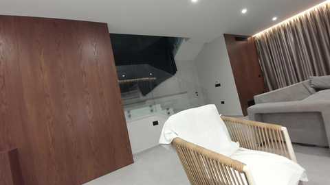 Video of a modern, minimalist living room with light wood furniture, white cushions, and beige curtains. The room features a staircase with a glass railing and a sleek, contemporary design.