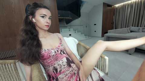 Video of a slender, fair-skinned woman with long, wavy brown hair, wearing a pink floral dress, sitting on a beige chair in a modern, minimalist living room with wooden panels and a white staircase.
