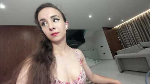 Video of a young woman with long brown hair, wearing a floral-patterned spaghetti-strap top, sitting in a modern, minimalist living room with white walls, wooden paneling, and a gray sofa.