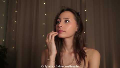 Media: Video of a young Asian woman with long dark hair, wearing a white top, looking thoughtful against a soft, dimly-lit curtain backdrop with twinkling fairy lights.
