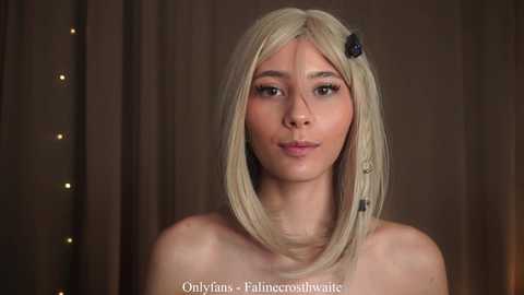 Video of a young, fair-skinned woman with straight, platinum blonde hair, adorned with a black hair clip, wearing minimal makeup. She stands against a brown curtain backdrop.