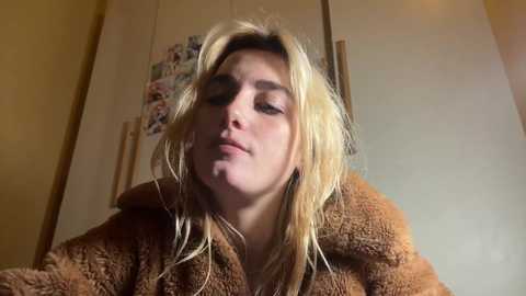 Video of a blonde woman with messy hair, wearing a brown fuzzy coat, leaning forward, indoors with white cabinets and a colorful magnet board in the background.