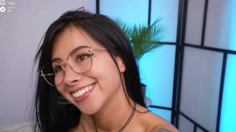 Video of a young woman with long black hair, wearing glasses, smiling, in a modern room with a blue-lit partition and green plant.