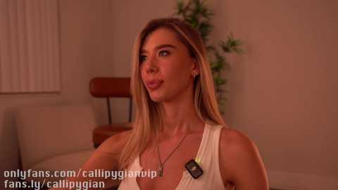 Media: Video of a blonde woman in a white tank top, sitting indoors with a potted plant behind her, looking thoughtful.