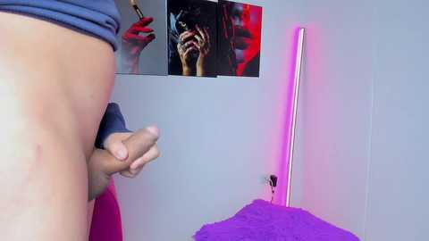 A video shows a man, nude from the waist down, holding an erect penis, standing in a room with a purple bed, wall-mounted artwork, and a neon pink light.