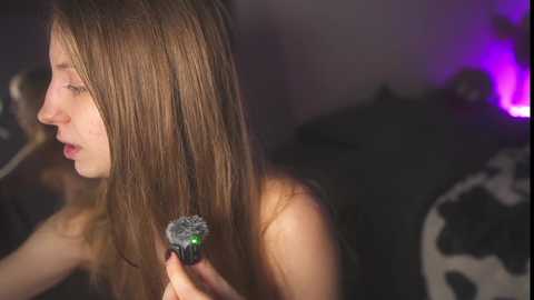 Video of a young woman with straight, long brown hair, using a hairbrush, indoors, with a dimly lit background featuring a cow print pillow and purple lighting.