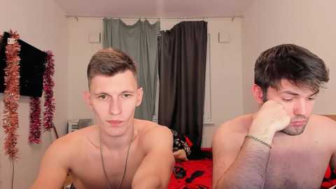 Video of two shirtless young men, one with short hair and a necklace, the other with dark hair and a bracelet, in a bedroom with red bedding, Christmas decorations, and a shower curtain.