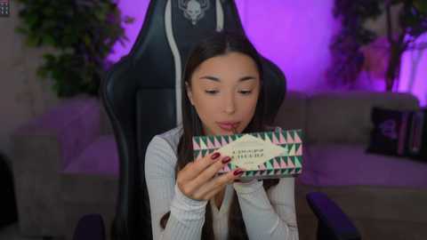 Video of a young woman with long brown hair, wearing a white sweater, holding a green and white box with pink geometric patterns, seated on a black gaming chair under purple lighting in a modern living room.