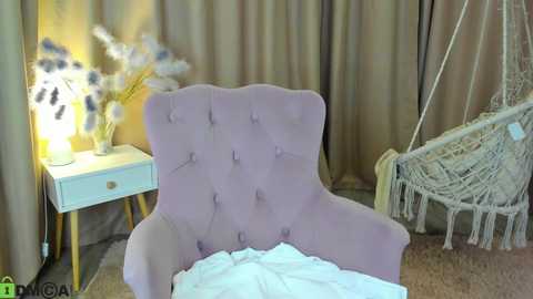 Video of a pastel purple velvet armchair with tufted buttons, next to a white side table with a lamp and floral arrangement. Behind, a beige curtain and a hanging white macram\u00e9 chair.
