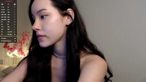 A video of a young woman with pale skin and long dark hair, wearing a thin silver necklace, looking contemplative against a plain gray background.
