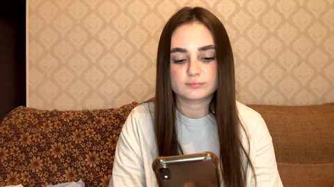 Video of a young Caucasian woman with long brown hair, wearing a white shirt, looking at a smartphone, sitting on a brown sofa with floral-patterned cushions. The background features a beige wallpaper with a diamond pattern.