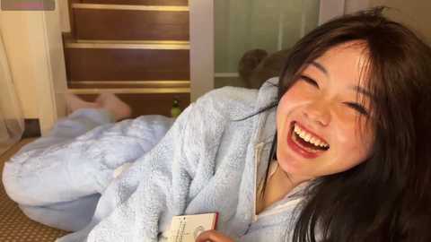 Video of an Asian woman with long black hair, smiling broadly, wearing a light blue, fuzzy bathrobe, holding a book, sitting on a beige carpeted floor, in a cozy indoor setting.