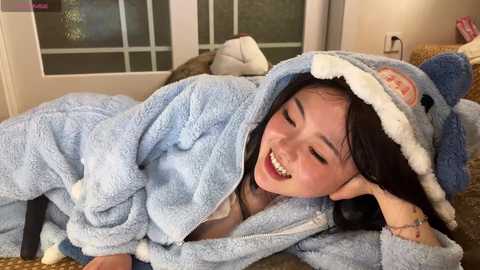Video of an Asian woman with long black hair, smiling, wearing a light blue, plush shark-themed robe, lying on a beige carpeted floor beside a window with white frames.