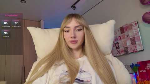 Video of a young, fair-skinned woman with long blonde hair, wearing a white graphic t-shirt, sitting on a cream-colored couch, surrounded by a cozy, modern bedroom with a pink wall, a TV, and a stuffed animal.