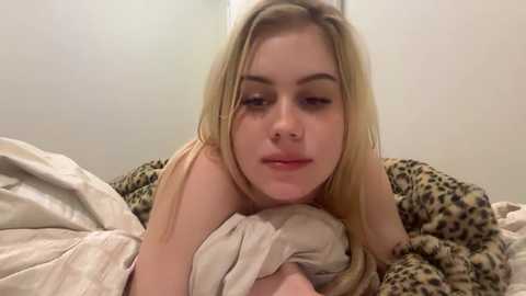 Video of a young, fair-skinned, blonde woman with straight hair, lying on a bed with beige and leopard-print sheets, looking down thoughtfully.