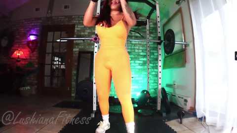 Video of a woman doing a burpee in a gym with a brick wall, green lighting, and fitness equipment. She wears a tight, yellow fitness outfit.