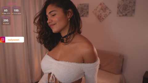 Media: Video of a smiling, light-skinned woman with wavy dark hair, wearing a white off-shoulder top, standing in a softly lit bedroom.