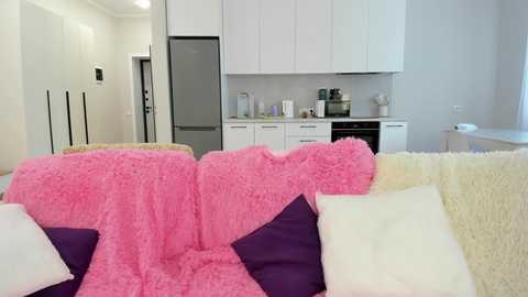 Video of a modern, minimalist kitchen with white cabinets and stainless steel appliances. In the foreground, a plush, pink, faux fur throw covers a white couch with purple and white pillows.