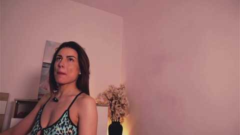 Video of a young woman with shoulder-length brown hair, wearing a leopard-print bra, standing against a beige wall with a floral painting and a vase of fake flowers on a table.