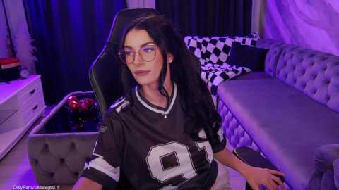 Video of a young woman with long black hair, wearing a black football jersey, glasses, and sitting on a grey couch. Background features purple lighting, checkered pillows, and a white dresser.