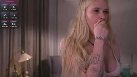 Video of a blonde woman with long hair, tattoos, and a thoughtful expression, holding her finger to her lips in a dimly lit room with a bed, lamp, and curtains.