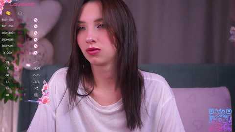 A video of a young Asian woman with long, straight black hair, wearing a white t-shirt, in a cozy room with pink flowers and a pink pillow.