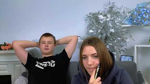 Media: Video of a young man in a black t-shirt, eyes closed, and a young woman with brown hair, wearing headphones, sitting on a gray couch. Background features a white fireplace, silver tinsel, and a vase with flowers.