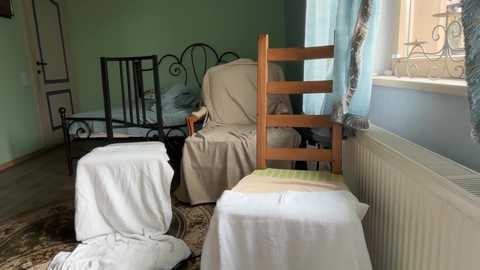Video of a cozy, cluttered bedroom with light green walls, a wooden chair, and a bed covered in white sheets, partially hidden by a beige curtain.