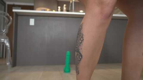 A video of a woman's leg with intricate tattoo on the inner thigh, standing in a modern kitchen with a green dildo on the floor.