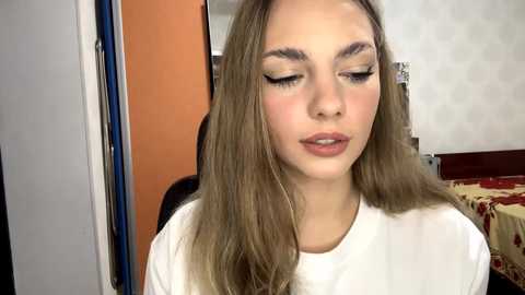 Video of a young Caucasian woman with long, straight, light brown hair, wearing white clothing, with subtle makeup, including winged eyeliner and nude lipstick, in a simple bedroom setting.