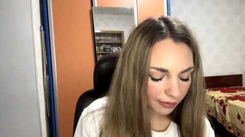 Video of a young woman with long, straight, light brown hair, wearing white clothing, sitting in a chair, looking down. Background includes a mirror, orange wall, and a bed with a red and beige patterned blanket.