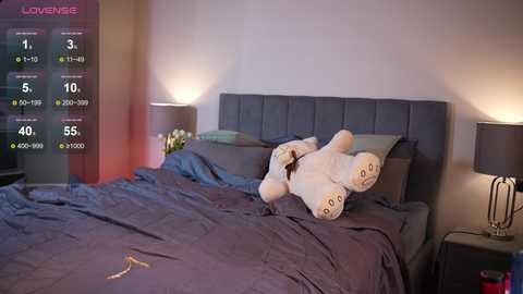 Media: A video of a modern, cozy bedroom with a plush, white teddy bear on a purple bedspread, beside a gray headboard, under soft, ambient lighting.