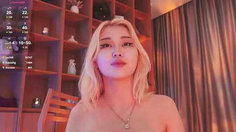 A video of a young, fair-skinned woman with blonde hair, wearing a necklace, sitting topless in a cozy room with wooden shelves and drapes.