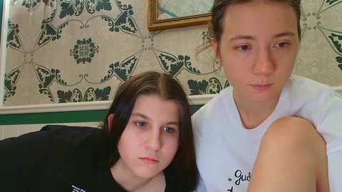 Video of two young girls with pale skin and straight brown hair, one in a black top and the other in a white tank, sitting in a room with green and white patterned wallpaper and a framed picture on the wall.