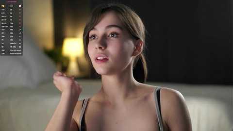 Media: Video of a young woman with light skin and brown hair, wearing a spaghetti-strap top, raising her right hand in a thoughtful pose. Background features a dimly lit room with a table lamp and blurred furniture.