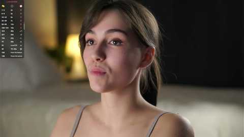 Video of a young woman with light skin and dark hair, wearing a light grey tank top, in a dimly lit room with blurred background elements.