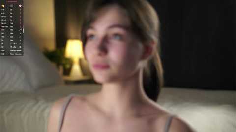 Media: Video of a young woman with fair skin, light brown hair, and blue eyes, wearing a light-colored top. Background shows a dimly lit bedroom with a bed, lamp, and green foliage.