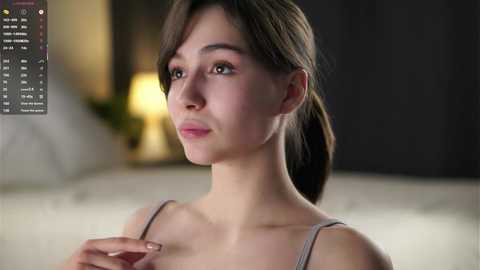 Media: Video of a young Asian woman with light skin, straight brown hair, and light makeup, wearing a grey spaghetti-strap top. She gazes thoughtfully to the left. Background shows blurred modern decor with a lamp.