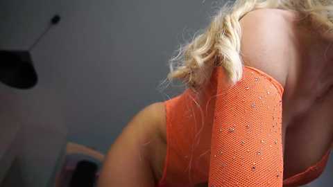 Video of a blonde woman with light skin wearing a bright orange, sequined, mesh sleeveless top, leaning forward, with a blurred, indistinct background.