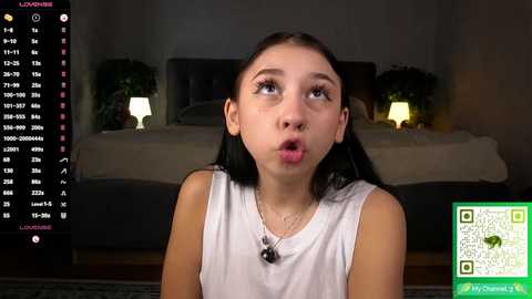 A video of a young Asian girl with straight black hair, wearing a white tank top, looking surprised in a dimly lit bedroom with a bed and green QR code.